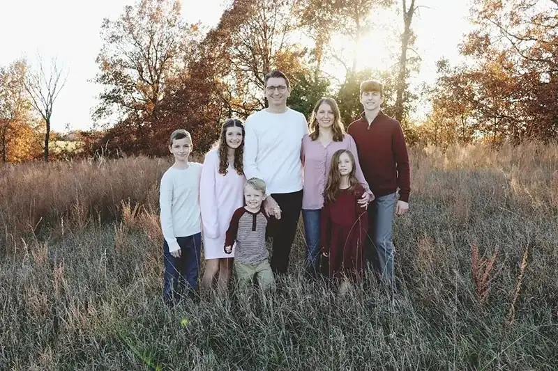 Family of six standing together in a grassy field during autumn with trees and warm sunlight in the background.